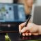 Close-up of a person using a stylus pen on a digital pad with a blurred laptop background.